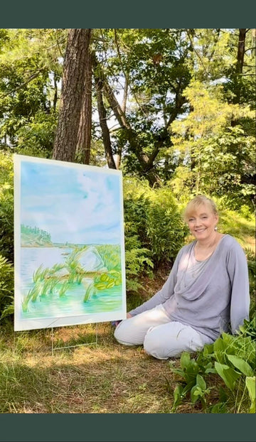 Pamela Hallock seated outdoors beside an original Georgian Bay watercolor painting, surrounded by summer trees and greenery.