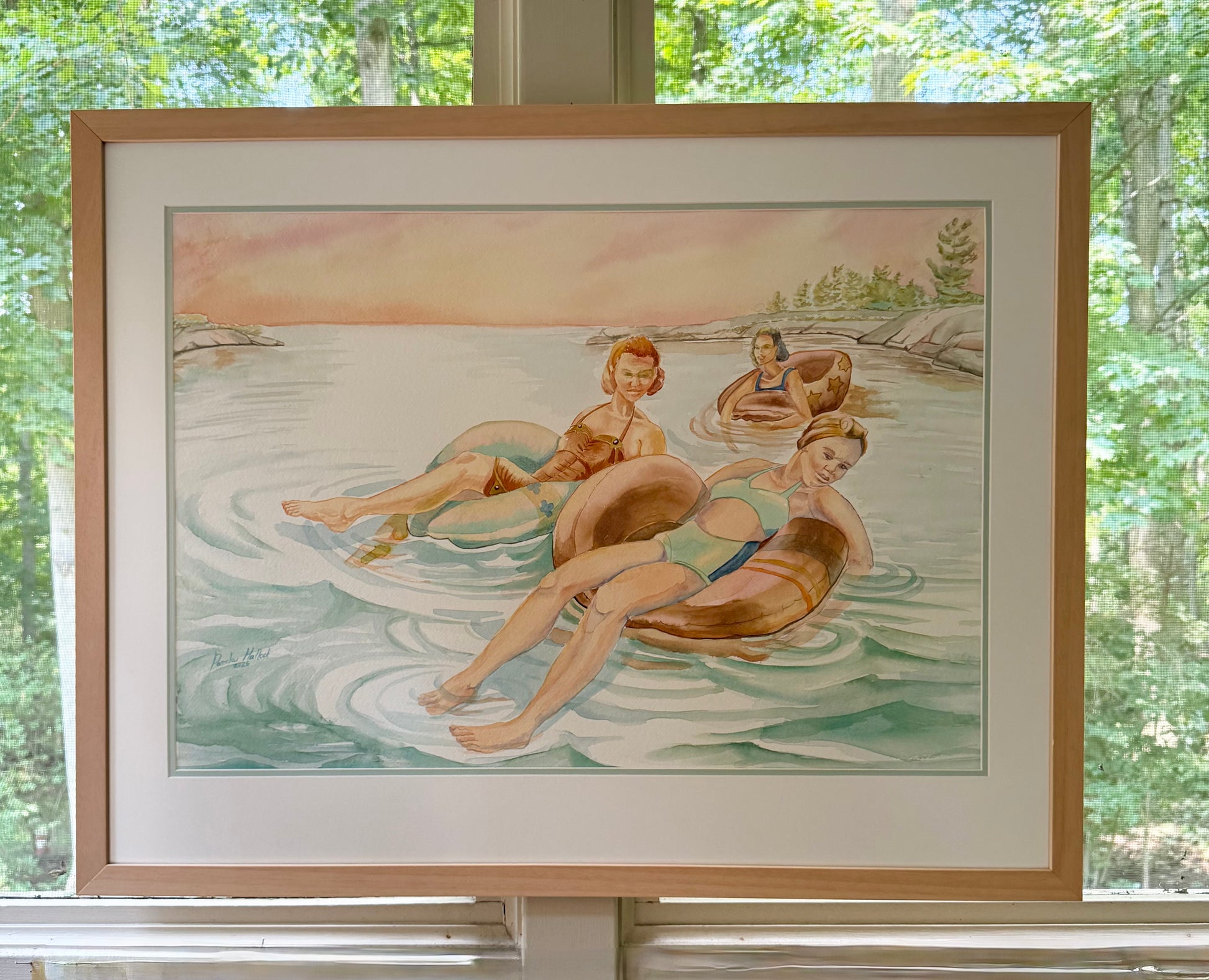 Framed original figurative watercolor painting "Pink Skies" by Pamela Hallock displayed in natural light, depicting three women floating on inner tubes in the calm waters of Georgian Bay beneath a soft pink evening sky.