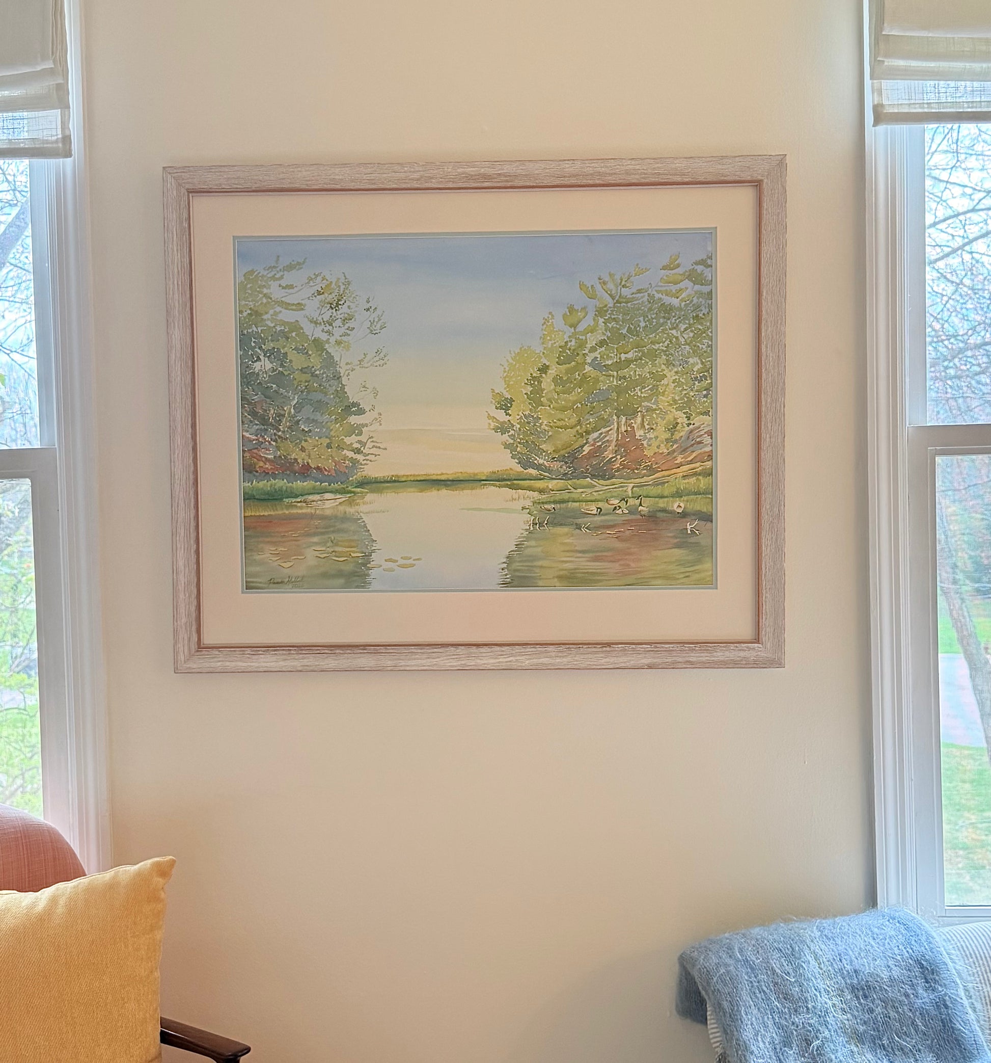 Framed original watercolor painting "Turtle Bay" displayed in a bright home setting, featuring peaceful sunrise reflections, calm water, water lilies, and shoreline pines inspired by Georgian Bay.