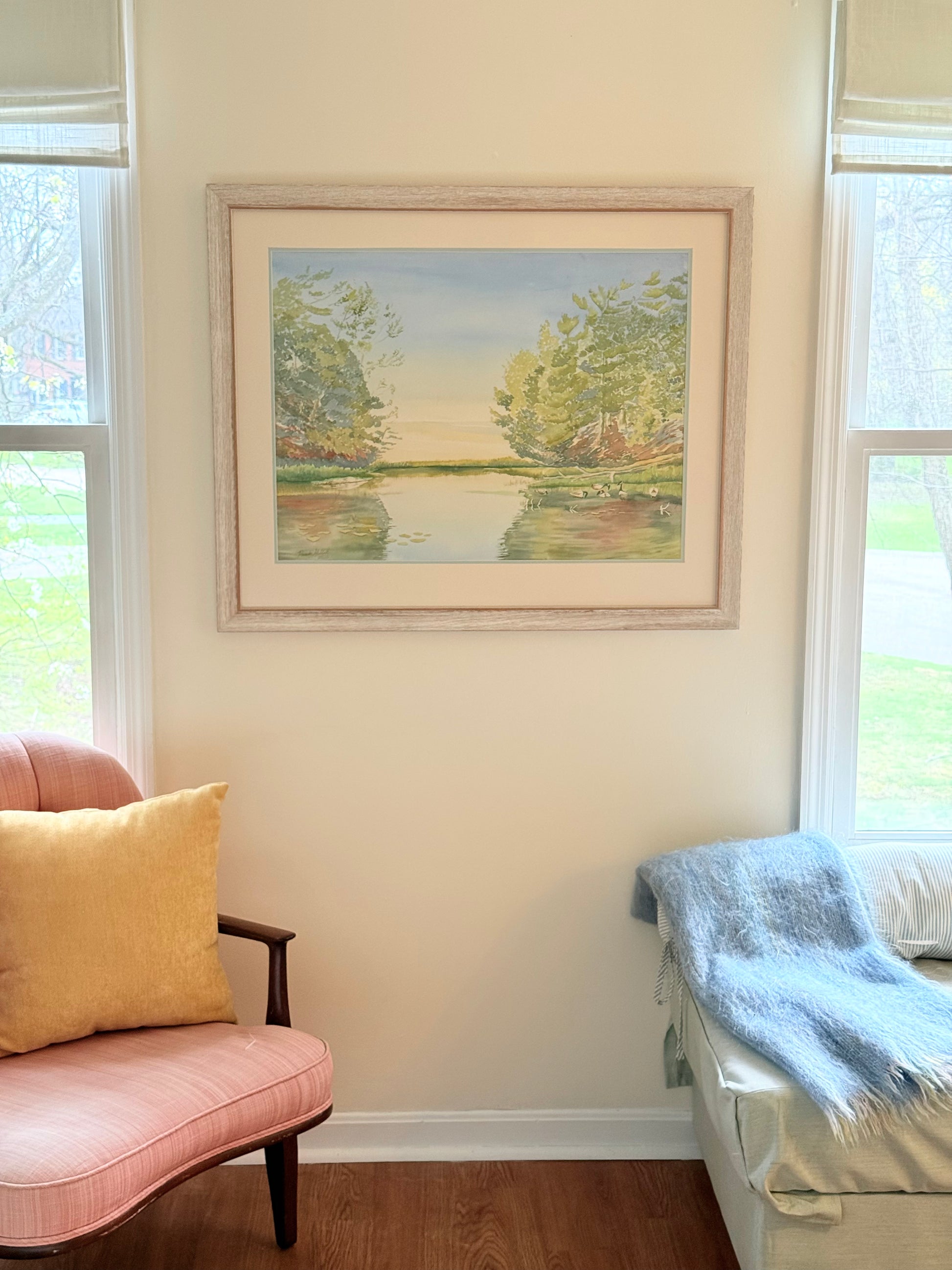 Framed original watercolor painting "Turtle Bay" displayed in a bright living room, featuring a peaceful sunrise over calm water with water lilies, shoreline pines, and soft reflections.